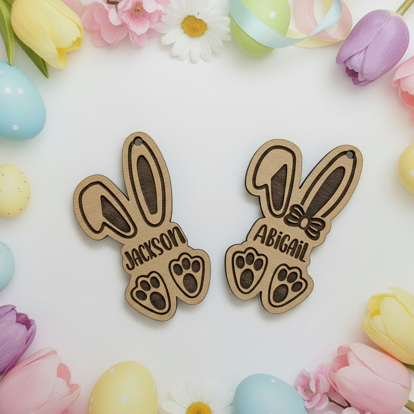 Two wooden bunny-shaped name tags with 'Jackson' and 'Abigail' engraved, surrounded by Easter eggs and flowers on a white background.